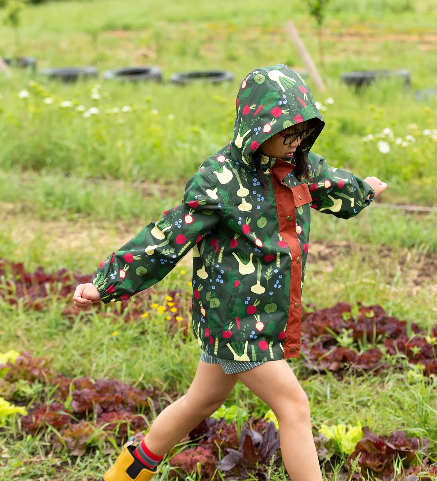 Child wearing the Little Green Radicals feast waterproof recycled kids raincoat with blue pinstripe shorts underneath with yellow wellington boots. 
