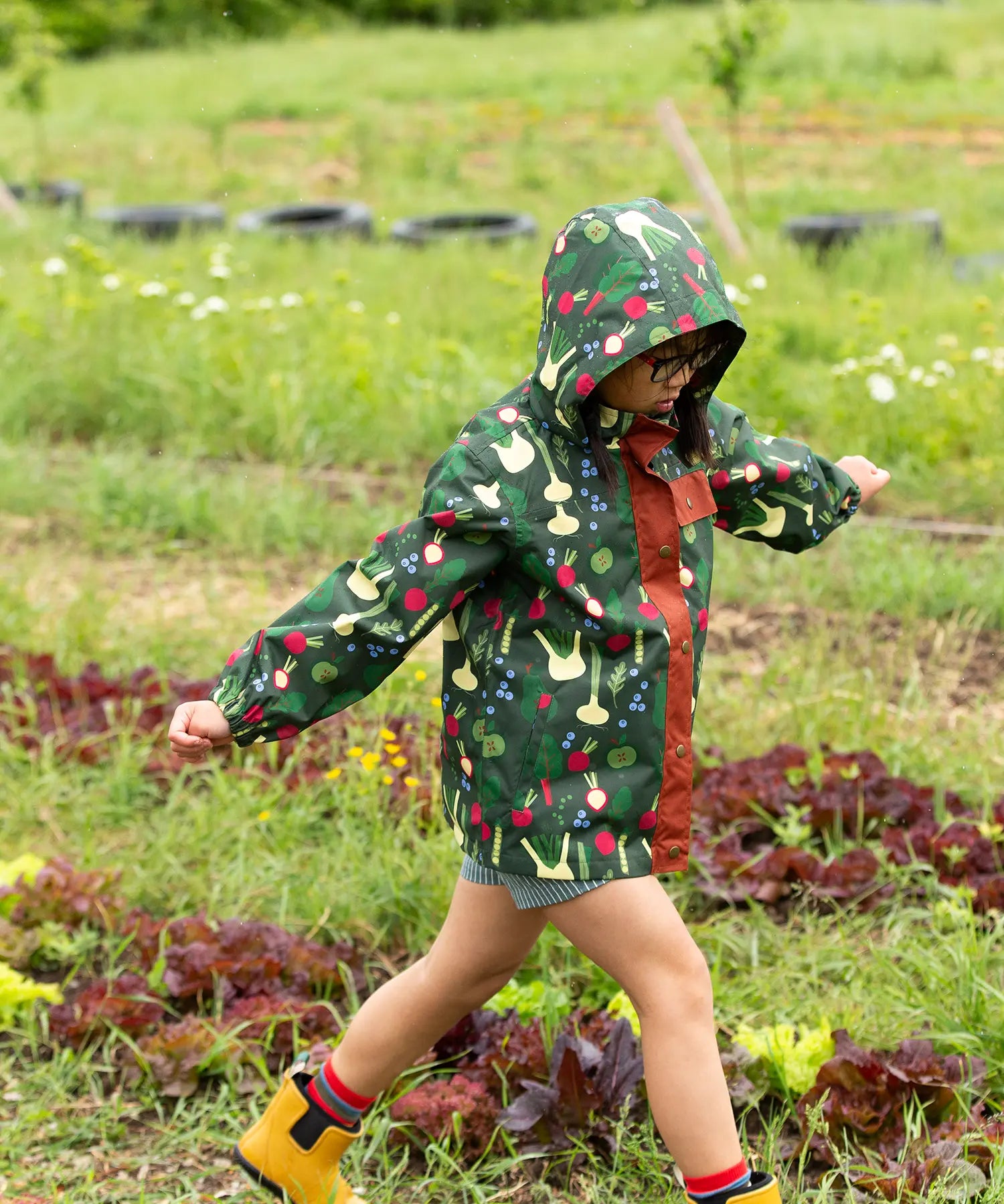 Child wearing the Little Green Radicals feast waterproof recycled kids raincoat with blue pinstripe shorts underneath with yellow wellington boots. 