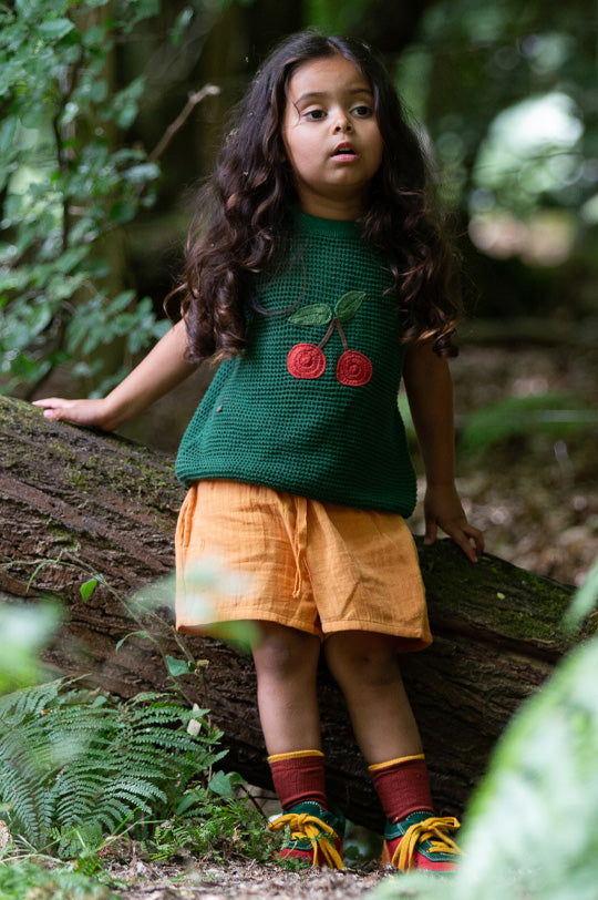 Child wearing Little Green Radicals Knitted Pullover in Green with large Cherry applique, made from organic cotton, paired with yellow muslin shorts.
