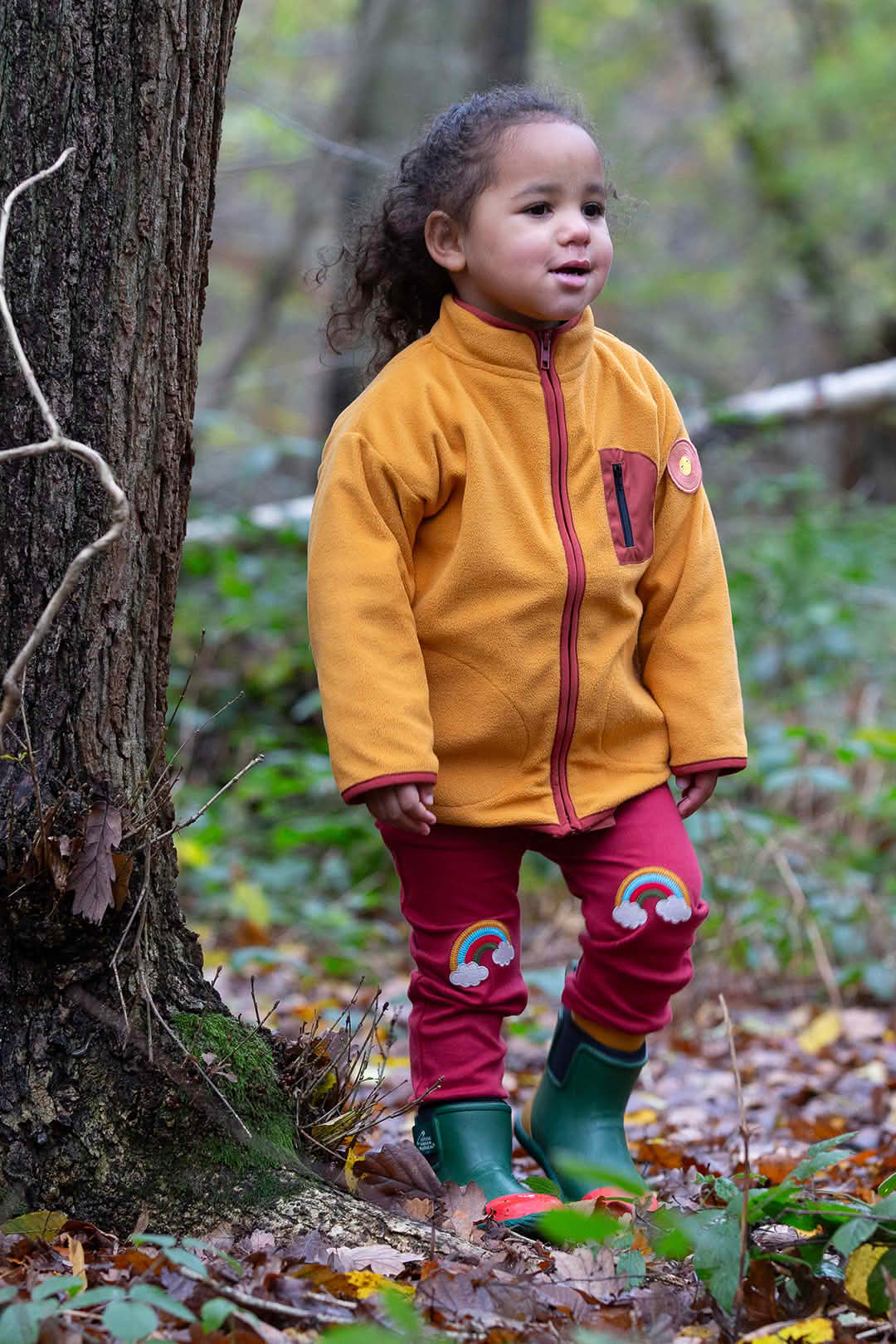 Babipur – Little Green Radicals Autumn Collection Fairtrade & GOTS Organic Kids Clothes. Child outdoors in forest wearing yellow organic fleece jacket and red trousers with rainbow knee patches, ethical and sustainable clothing at Babipur.