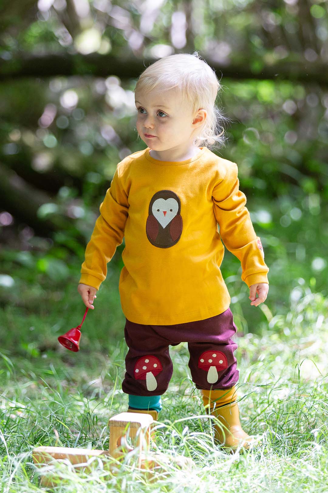 Child wearing  Little Green Radicals yellow shirt with a brown owl design, purple pants with red mushroom applique, and yellow fairtrade welly boots standing in a grassy area. This images is to represent the brand at babipur. 