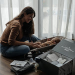 A person sits in front of their child which is in a baby boucer chair, with the Lola & Lykke Breastfeeding Starter Kit next to them