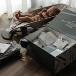 An open box showing the contents of the Lola & Lykke Breastfeeding Starter Kit. A baby is in the background in a baby bouncer