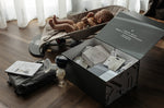 An open box showing the contents of the Lola & Lykke Breastfeeding Starter Kit. A baby is in the background in a baby bouncer