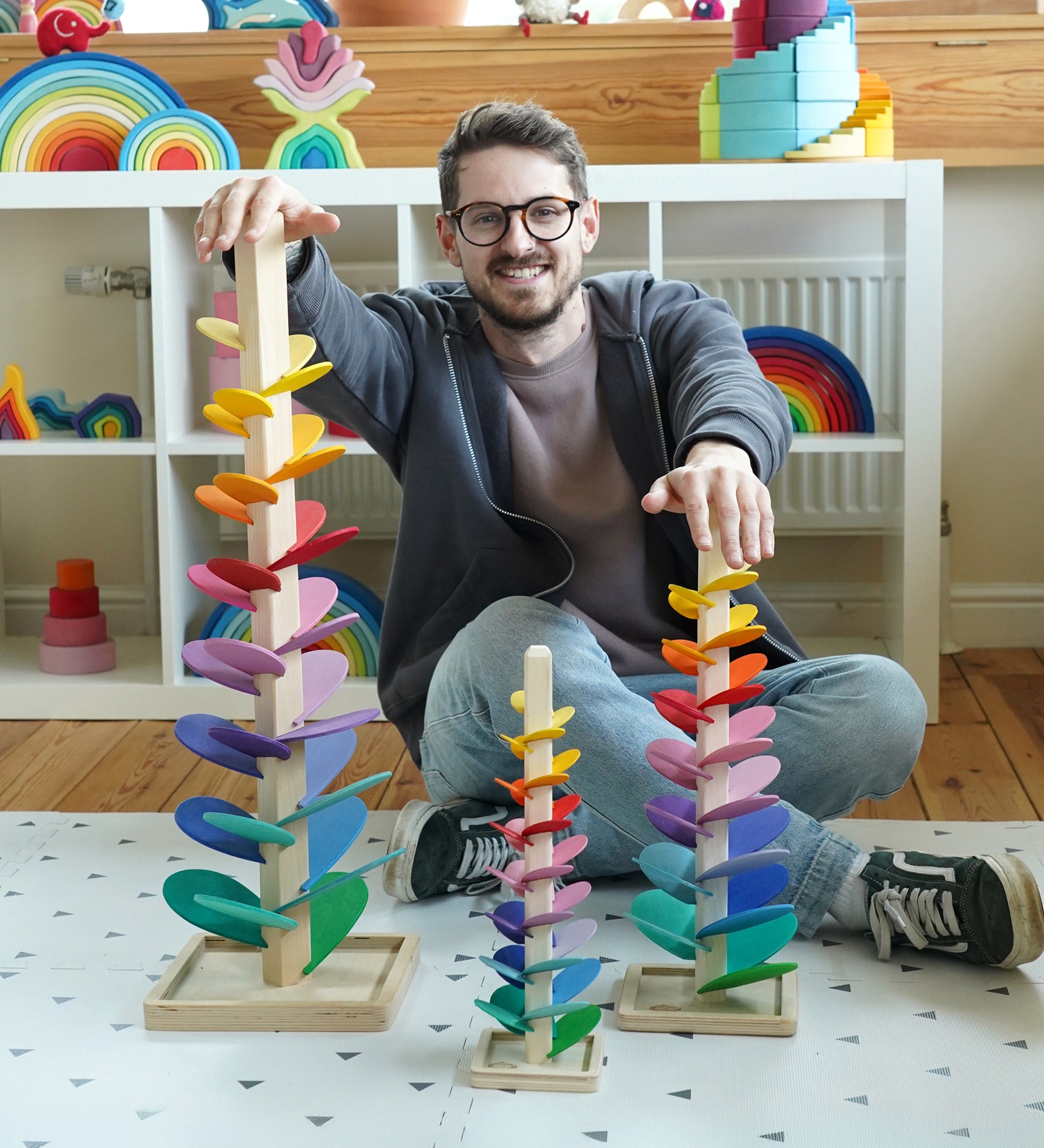Comparison of Magic Wood Marble trees: mini, small and large next to a man in the Babipur playroom. Satisfying wooden marble runs made of a tall stem, rainbow leaves and marbles.