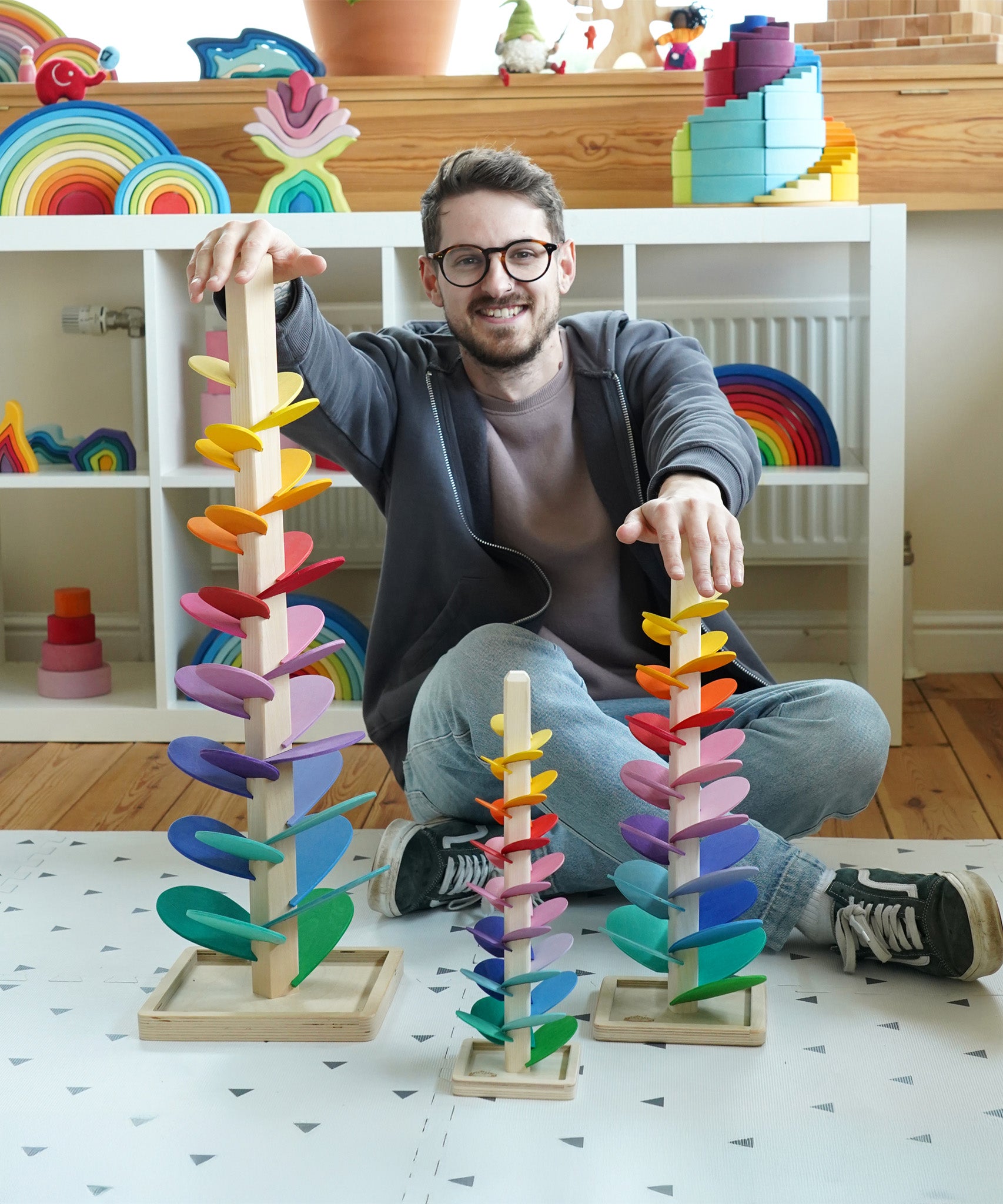 Comparison of Magic Wood Marble trees: mini, small and large next to a man in the Babipur playroom. Satisfying wooden marble runs made of a tall stem, rainbow leaves and marbles.