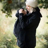 Side of mother wearing Mamalila's soft shell baby wearing allrounder fitted babywearing jacket in black with the hood covering baby's head