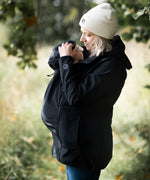 Side of mother wearing Mamalila's soft shell baby wearing allrounder fitted babywearing jacket in black with the hood covering baby's head