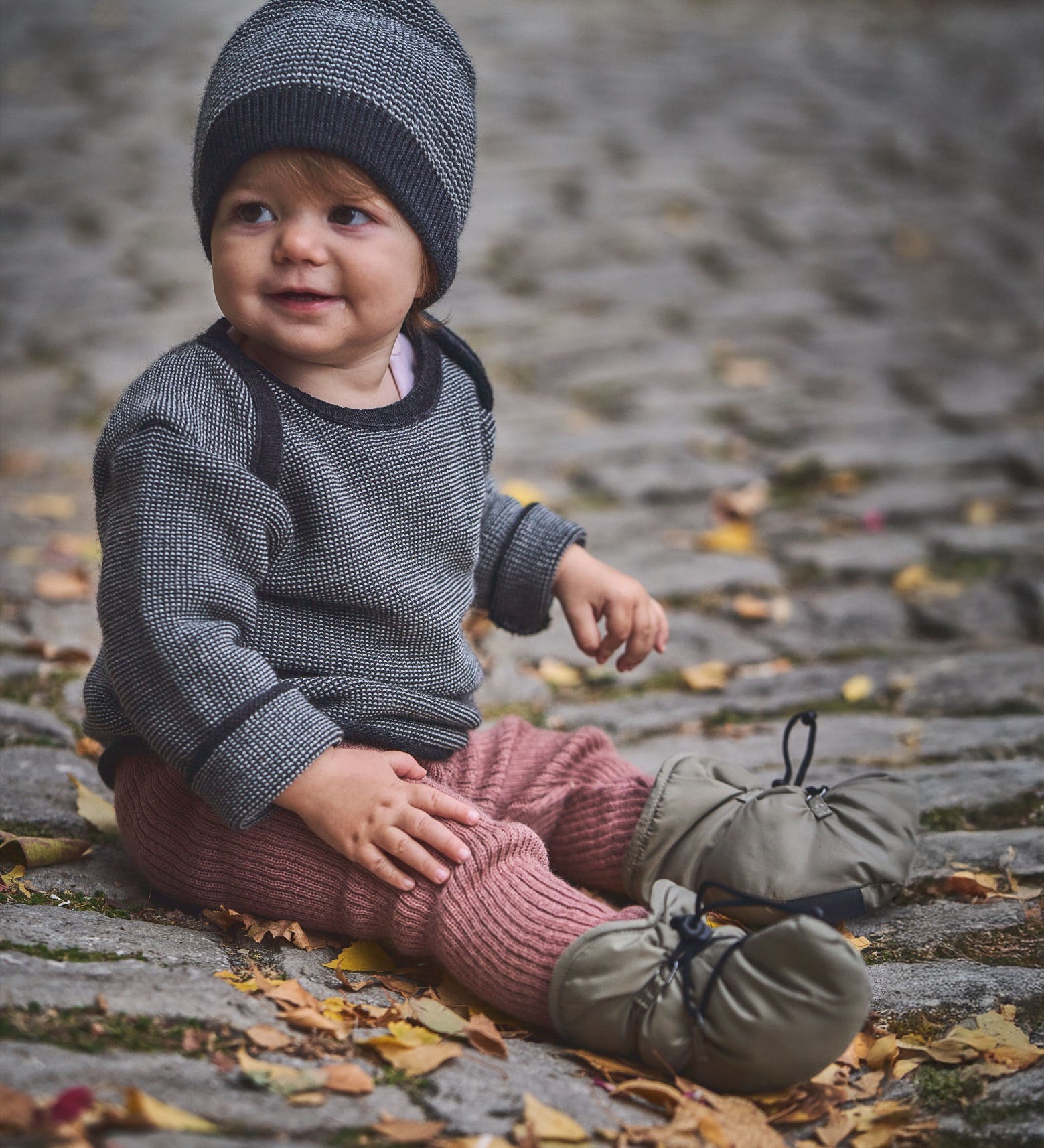 A child sitting on a cobbled surface and wearing the Mamalila quilted Berlin booties in Khaki available at Babipur.