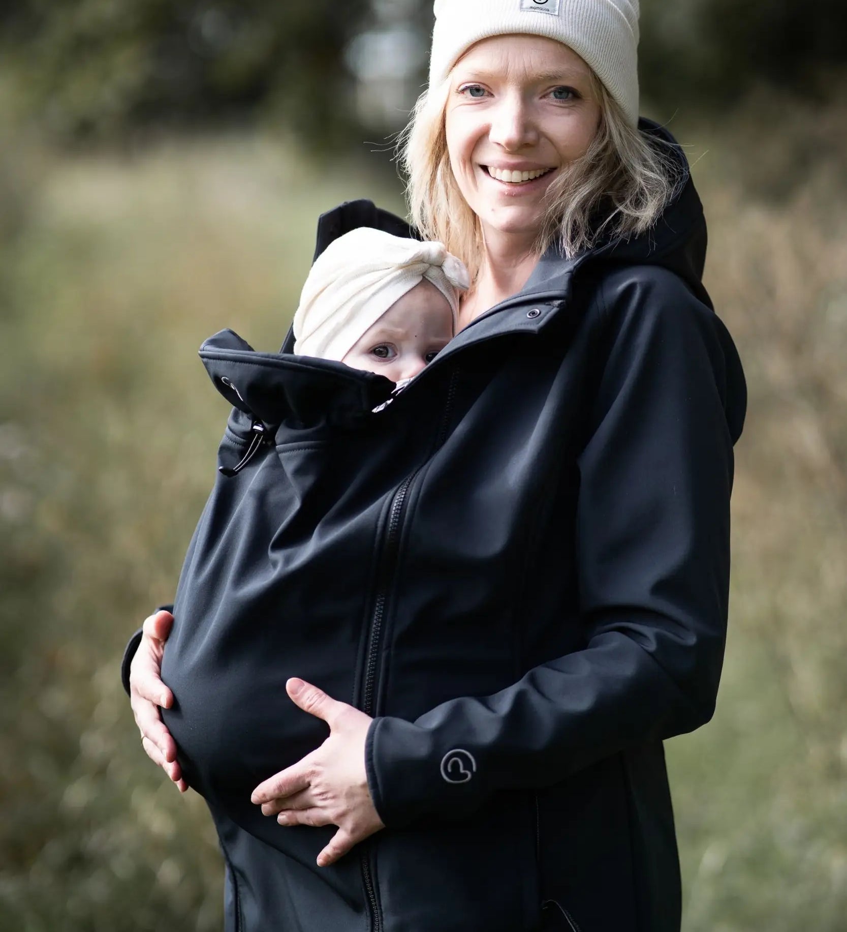 A mother wearing the Mamalila soft shell baby wearing allrounder fitted jacket in black carrying a baby on the front