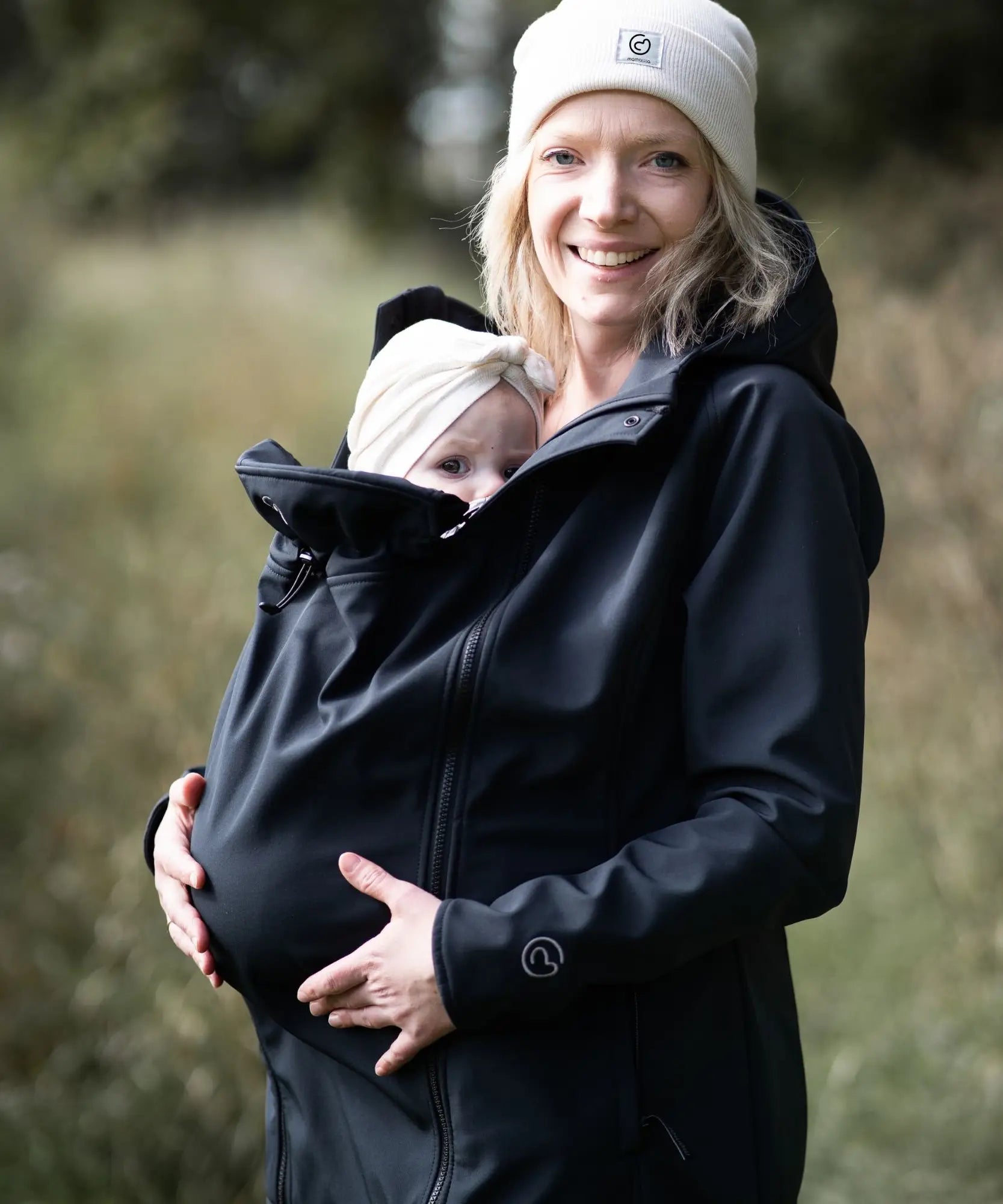 A mother wearing the Mamalila soft shell baby wearing allrounder fitted jacket in black carrying a baby on the front