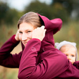 A mother wearing the Mamalila soft shell baby wearing allrounder fitted jacket in burgundy putting the hood on carrying a baby on the back