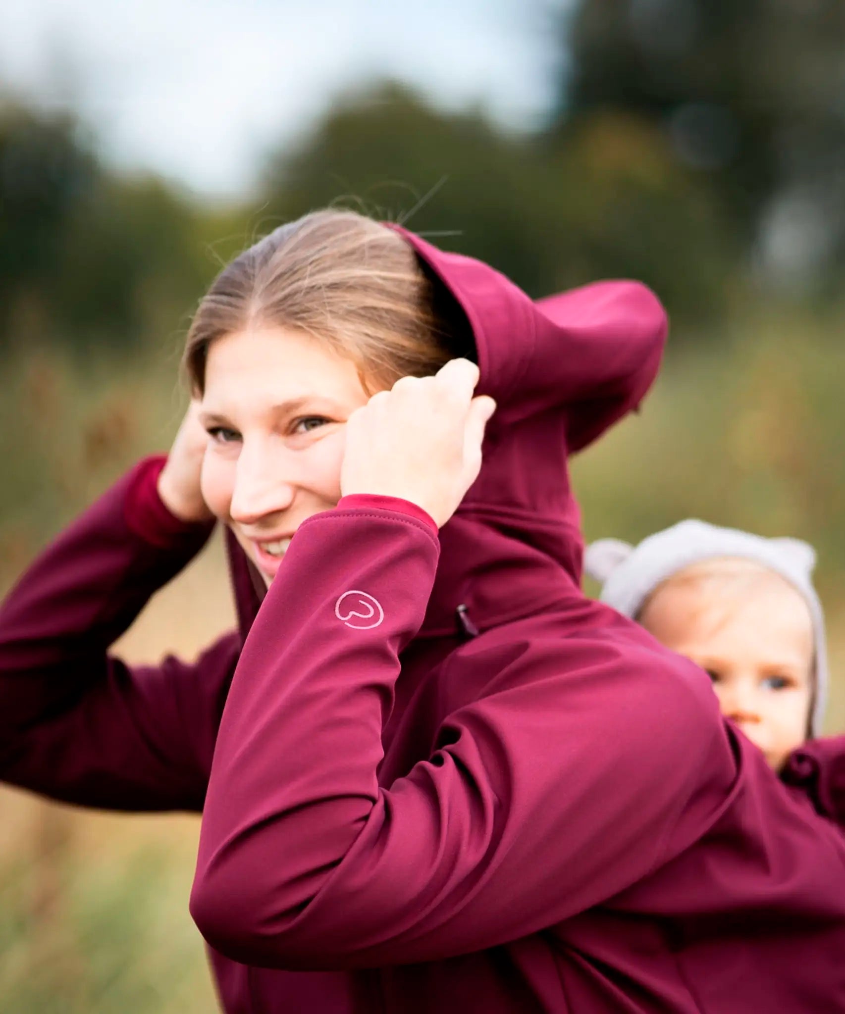A mother wearing the Mamalila soft shell baby wearing allrounder fitted jacket in burgundy putting the hood on carrying a baby on the back
