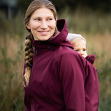 A mother wearing the Mamalila soft shell baby wearing allrounder fitted jacket in burgundy carrying a baby on the back