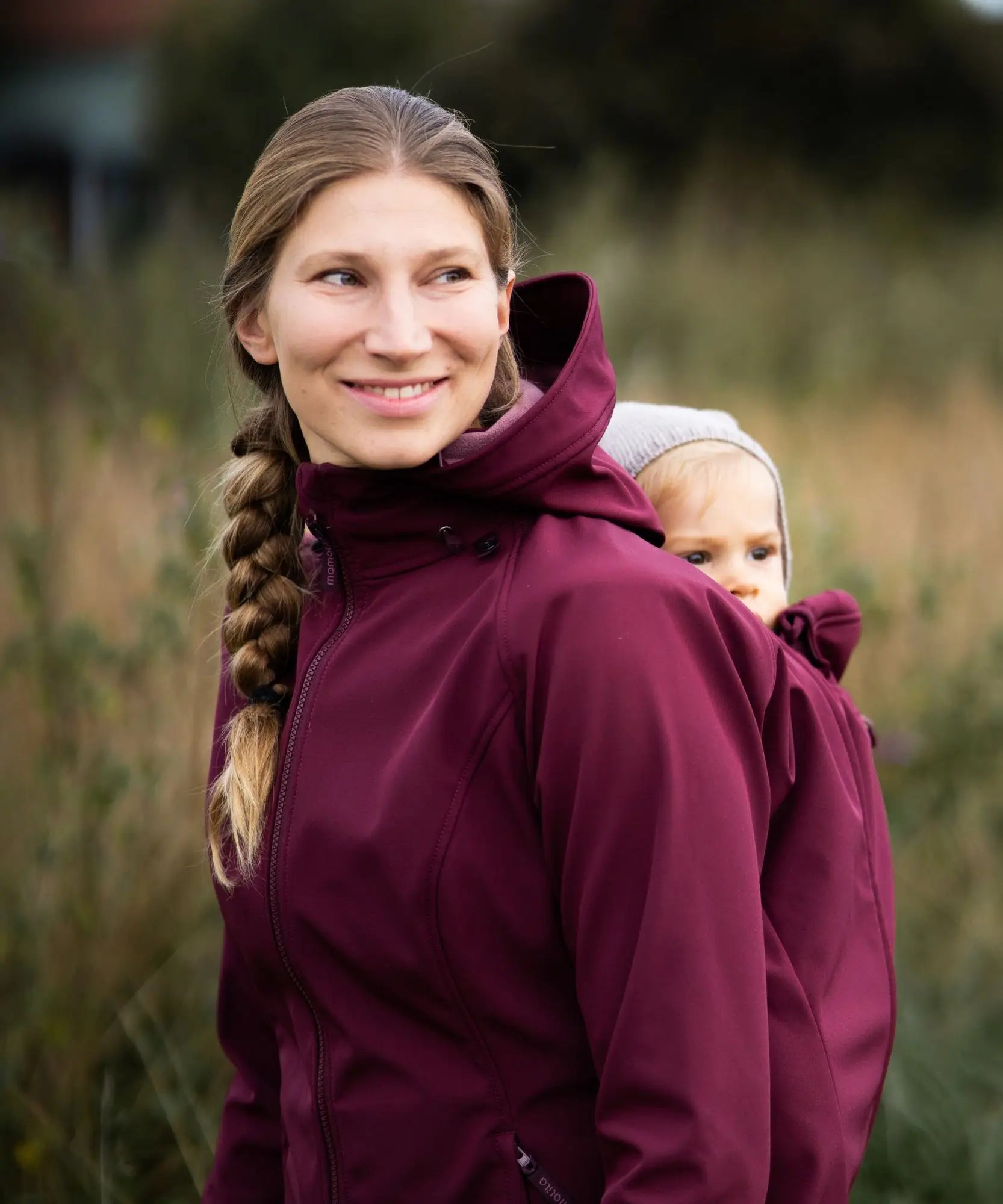 A mother wearing the Mamalila soft shell baby wearing allrounder fitted jacket in burgundy carrying a baby on the back