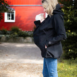 Side of mother wearing Mamalila's soft shell baby wearing allrounder fitted jacket in black carrying a baby on the front with a red house in the background.