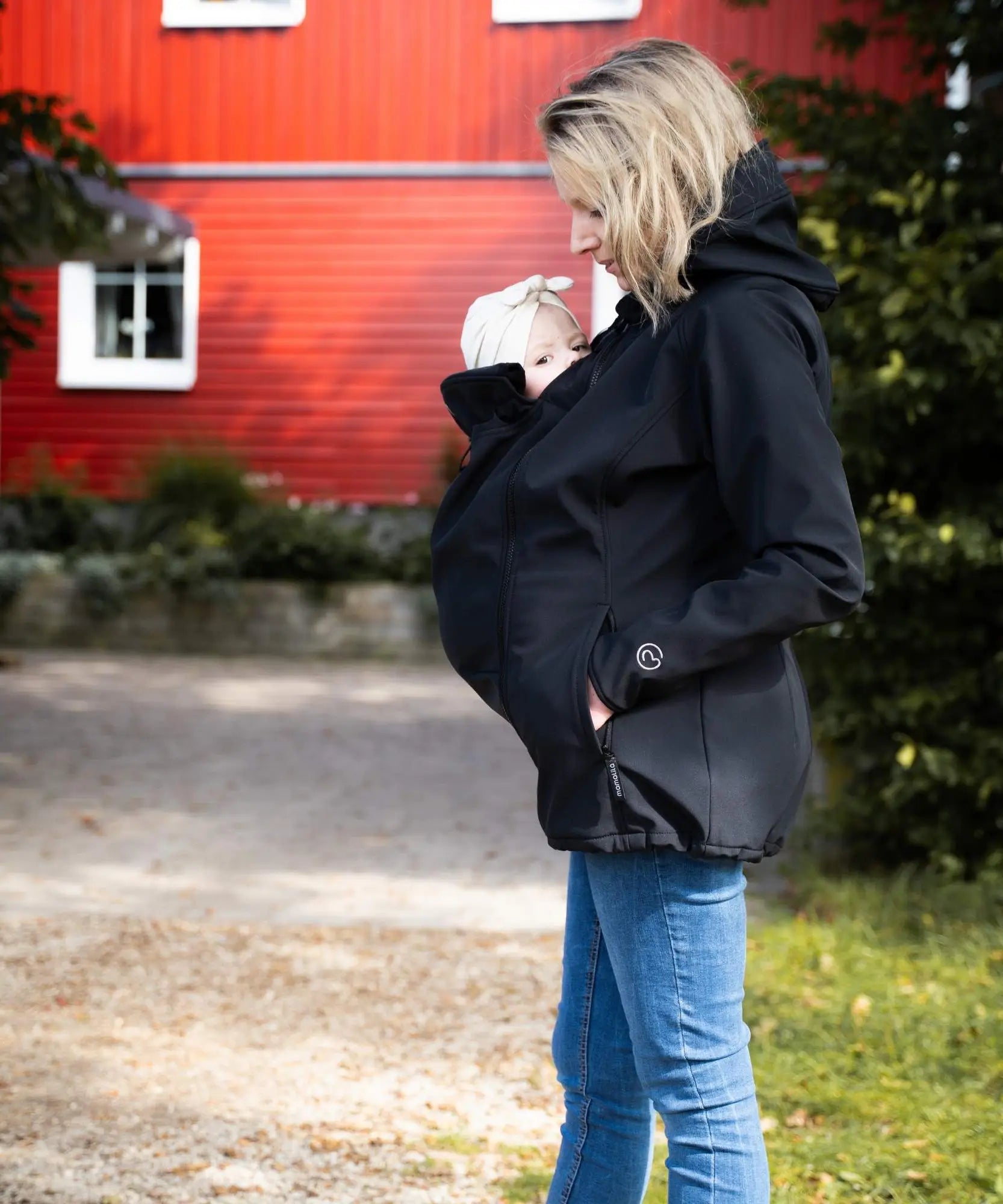 Side of mother wearing Mamalila's soft shell baby wearing allrounder fitted jacket in black carrying a baby on the front with a red house in the background.