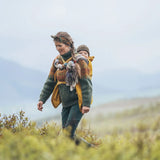 Woman carrying baby on the back with a yellow Mamalila universal softshell babywearing cover over the carrier with mountains in the background.