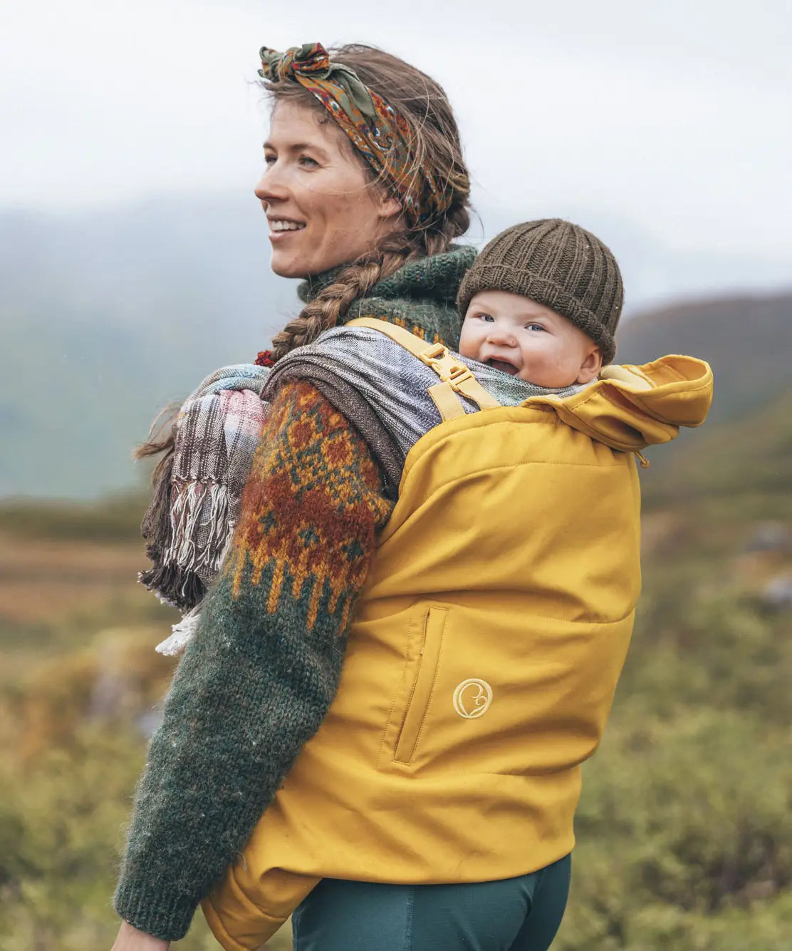 Woman with a baby in carrier with a yellow Mamalila universal softshell babywearing cover with mountains in the background.