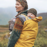 Woman with a baby in carrier with a yellow Mamalila universal softshell babywearing cover with mountains in the background.