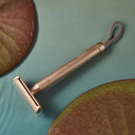 MÜHLE Companion Razor - Rose Gold, is sat on a blue table that looks like water, with two water lilies either side 