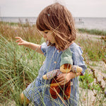 A child holding a Nanchen Forest Child Waldorf Doll in their arms, whilst walking along a grassy beach area