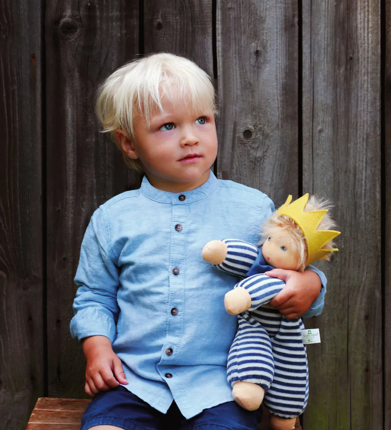 Child playing with Nanchen King Charles waldorf cuddle doll on a wooden bench doll has a blue and white outfit, yellow crown and blue scarf