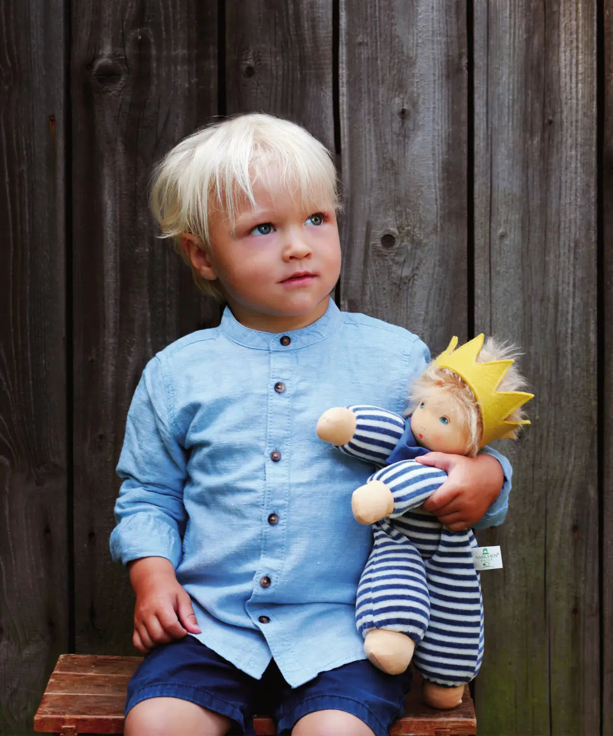 Child playing with Nanchen King Charles waldorf cuddle doll on a wooden bench doll has a blue and white outfit, yellow crown and blue scarf