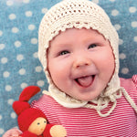A child is happily lying down on a light blue and white spot blanket, with a big smile on their face, and is wearing a crochet cream bonnet and red stripe top. The child is holding The Nanchen Waldorf Pimpel Doll in red