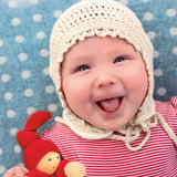 A child is happily lying down on a light blue and white spot blanket, with a big smile on their face, and is wearing a crochet cream bonnet and red stripe top. The child is holding The Nanchen Waldorf Pimpel Doll in red