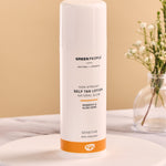 Green People Self Tan Lotion 150ml on a white wooden plate and a flower in a glass vase in the background