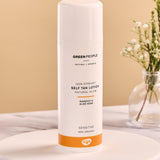 Green People Self Tan Lotion 150ml on a white wooden plate and a flower in a glass vase in the background