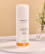Green People Self Tan Lotion 150ml on a white wooden plate and a flower in a glass vase in the background