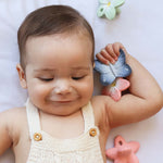 A child smiling and holding the Oli & Carol Natural Rubber Mini Teether - Blues The Butterfly in their hand. Around the child are other Oli & Carol natural rubber teethers