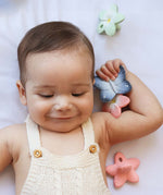 A child smiling and holding the Oli & Carol Natural Rubber Mini Teether - Blues The Butterfly in their hand. Around the child are other Oli & Carol natural rubber teethers