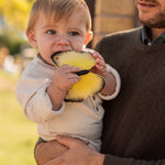 A child being held by an adult, chewing on the yellow Oli & Carol Natural Rubber Teether - Katia The Butterfly