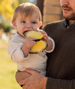 A child being held by an adult, chewing on the yellow Oli & Carol Natural Rubber Teether - Katia The Butterfly