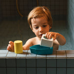 A child in a bath, playing with the blue Oli & Carol Boat Bath Toy, made from 100% Upcycled Natural Rubber. The child is playing the parts of the boat together