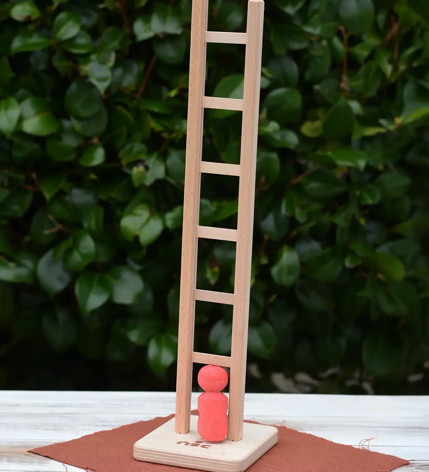 Nic climbing wooden ladder toy with a small red peg person standing on the base,  outdoors on a white tabletop with a leafy green hedge in the background.