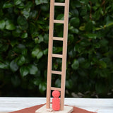 Nic climbing wooden ladder toy with a small red peg person standing on the base,  outdoors on a white tabletop with a leafy green hedge in the background.