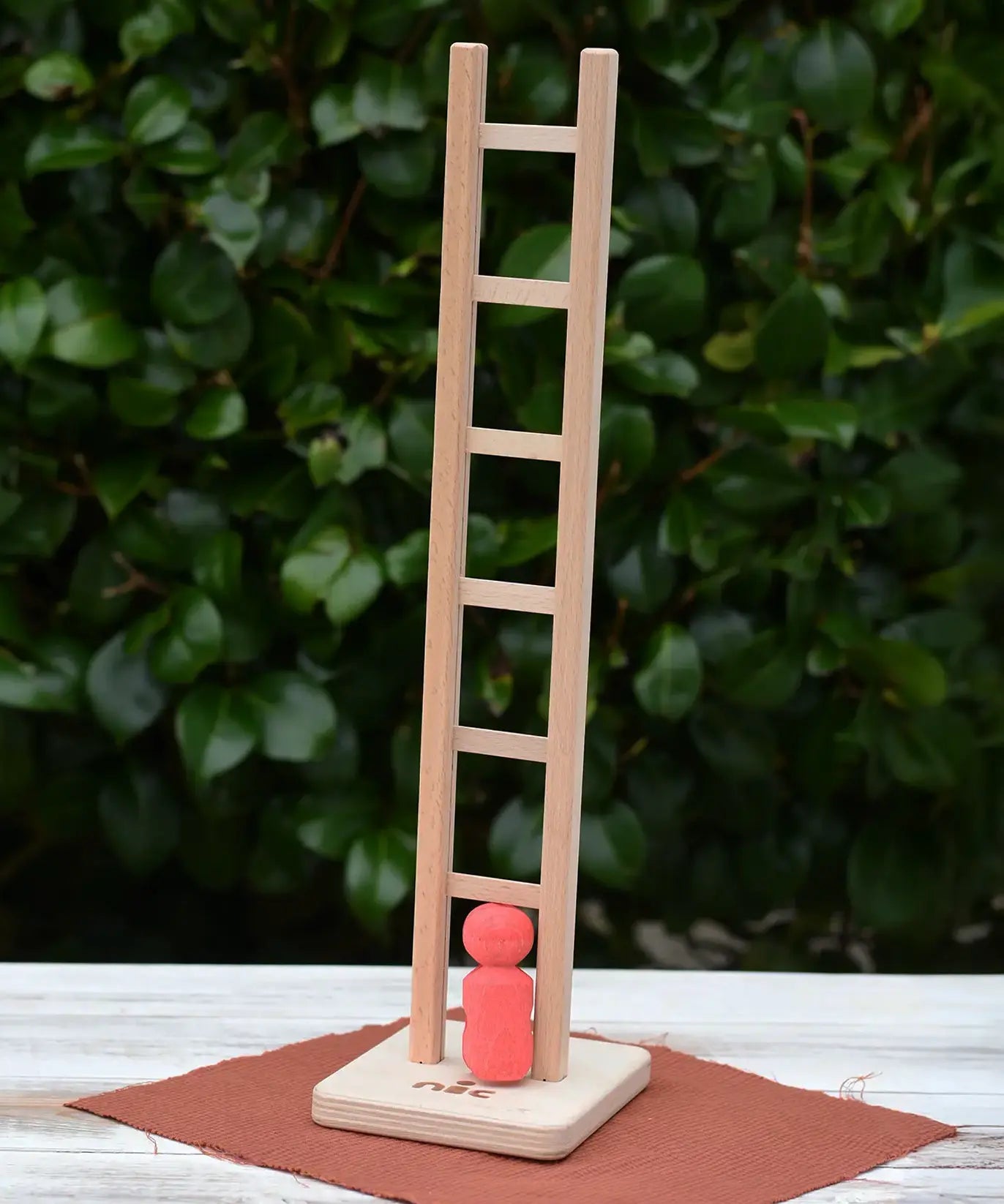 Nic climbing wooden ladder toy with a small red peg person standing on the base,  outdoors on a white tabletop with a leafy green hedge in the background.