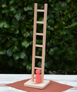 Nic climbing wooden ladder toy with a small red peg person standing on the base,  outdoors on a white tabletop with a leafy green hedge in the background.