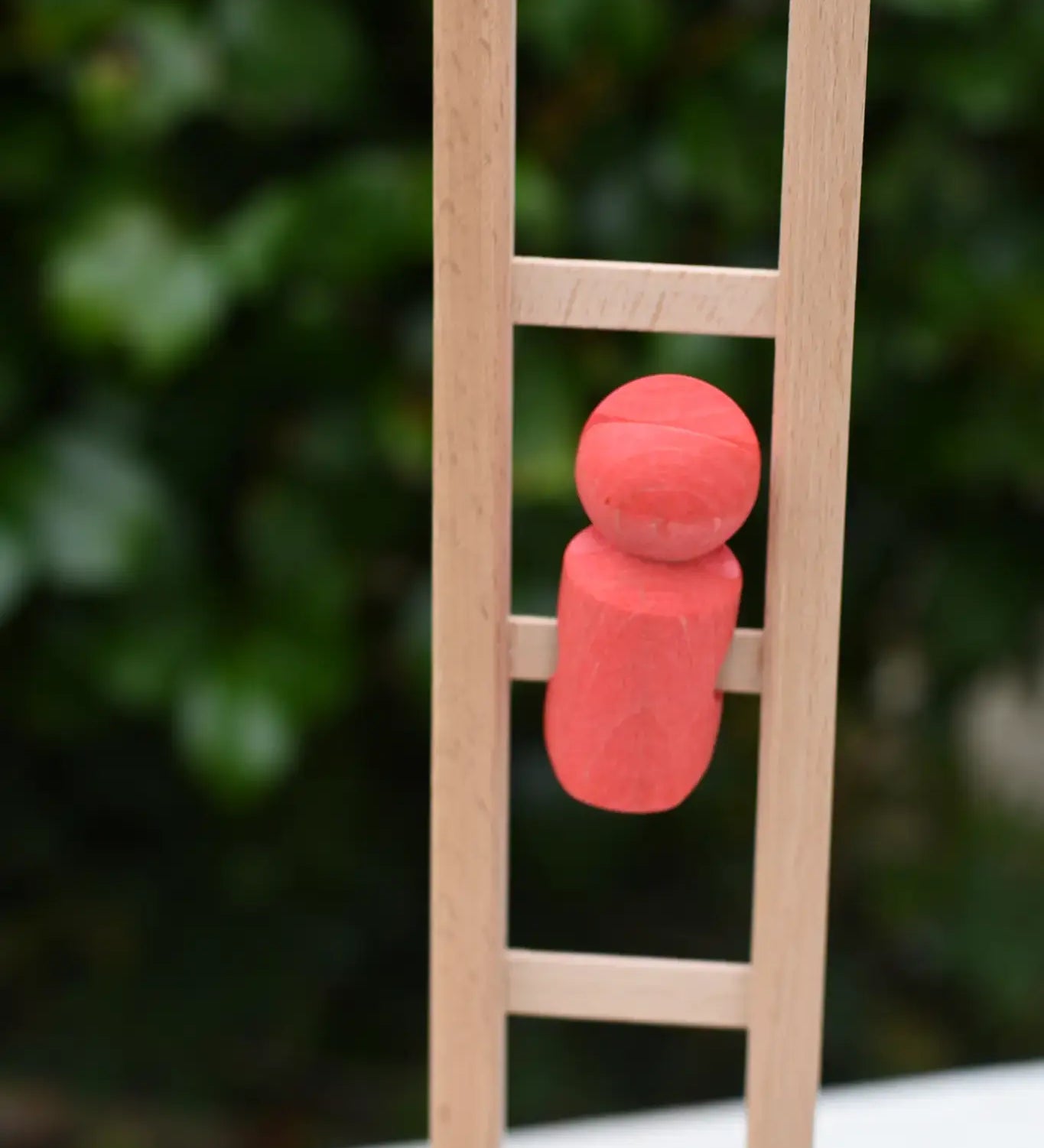 A small coral-red wooden toy figure from the Nic climbing wooden ladder toy stands on the light natural wood ladder against a blurred green outdoor background.