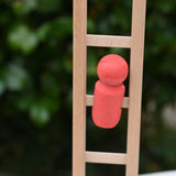 A small coral-red wooden toy figure from the Nic climbing wooden ladder toy stands on the light natural wood ladder against a blurred green outdoor background.