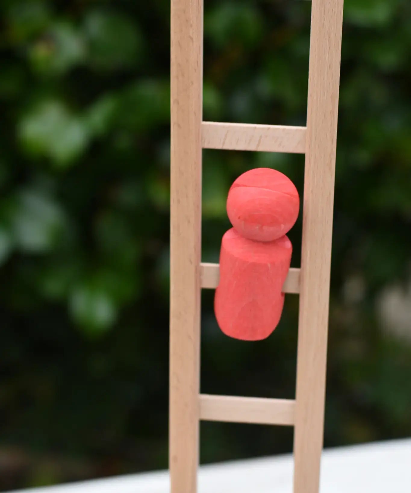 A small coral-red wooden toy figure from the Nic climbing wooden ladder toy stands on the light natural wood ladder against a blurred green outdoor background.