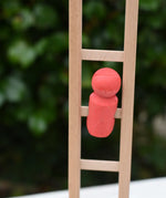 A small coral-red wooden toy figure from the Nic climbing wooden ladder toy stands on the light natural wood ladder against a blurred green outdoor background.