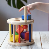 Nic rolling sorter wooden shape-sorting toy with colorful geometric blocks, displayed on a wooden table with a child's hand reaching in to touch the blue block on top.