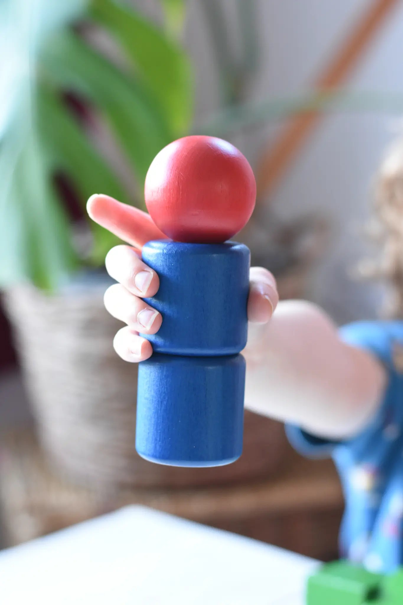 A child's hand holding a wooden toy peg dolls style figure with a red round head and blue stacked body parts, created using parts from the Nic screws and nuts colorful wooden toy threaded blocks and shapes set.