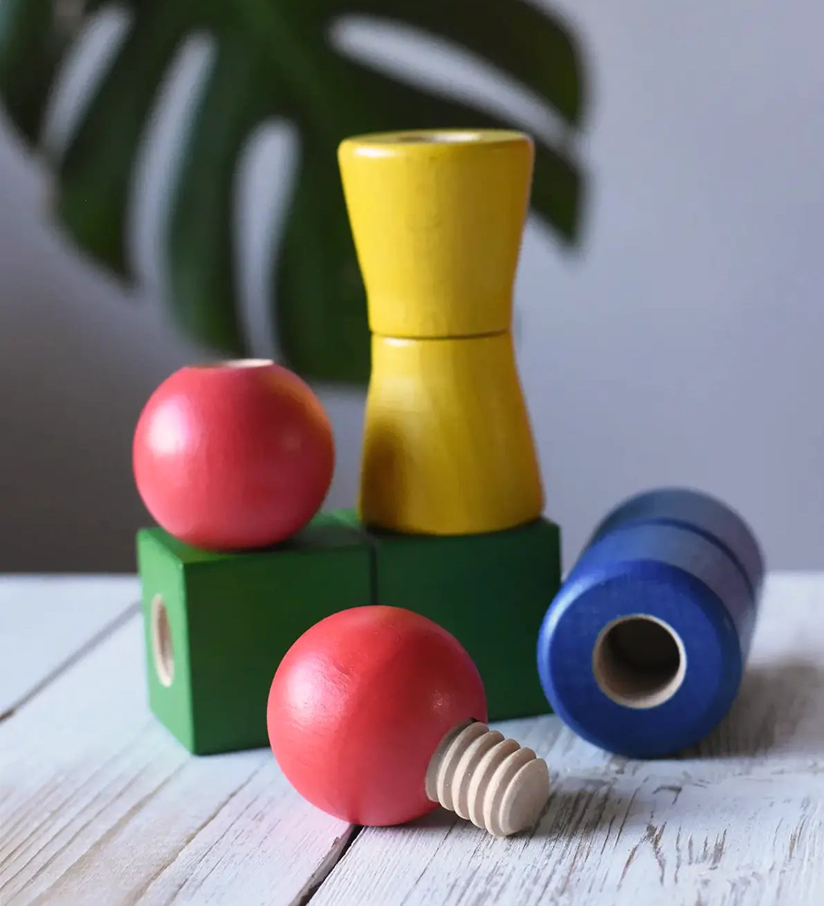 Nic screws and nuts colorful wooden toy threaded blocks and shapes arranged on a white wooden surface, including red spherical pieces, a green block, a yellow cone-shaped piece, and a blue cylindrical piece, with a blurred green leaf in the background.