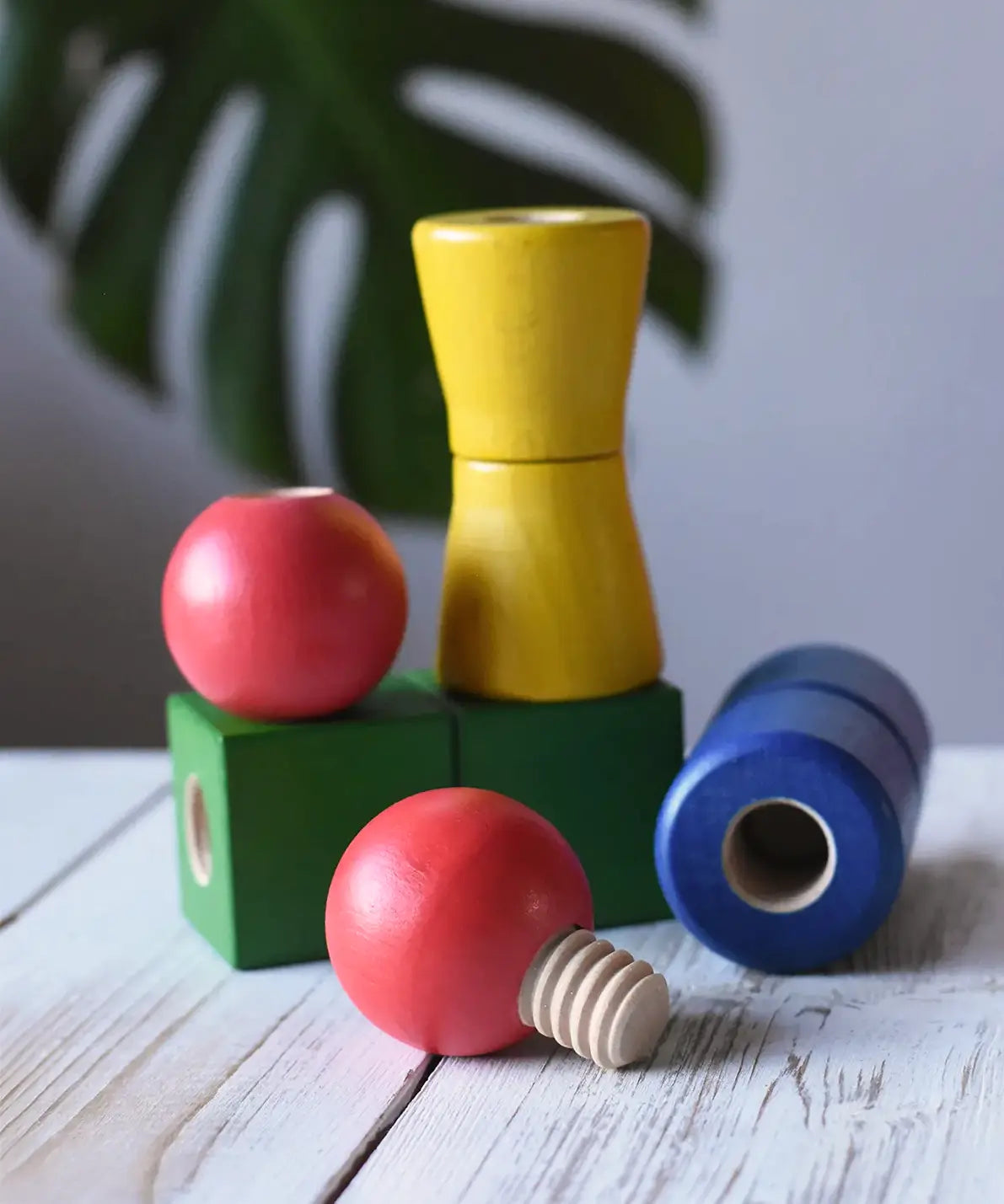 Nic screws and nuts colorful wooden toy threaded blocks and shapes arranged on a white wooden surface, including red spherical pieces, a green block, a yellow cone-shaped piece, and a blue cylindrical piece, with a blurred green leaf in the background.